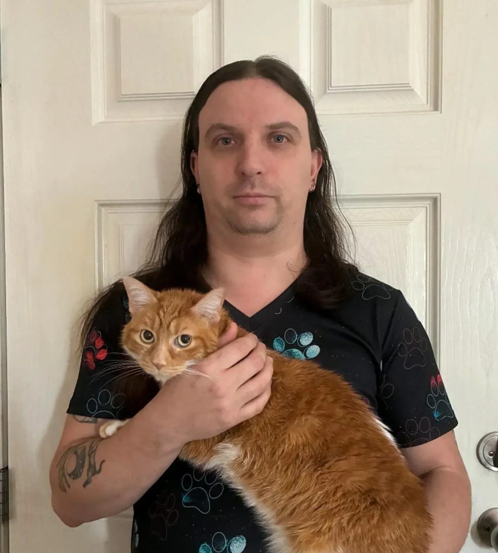 Man with long hair holding an orange cat in front of a white door