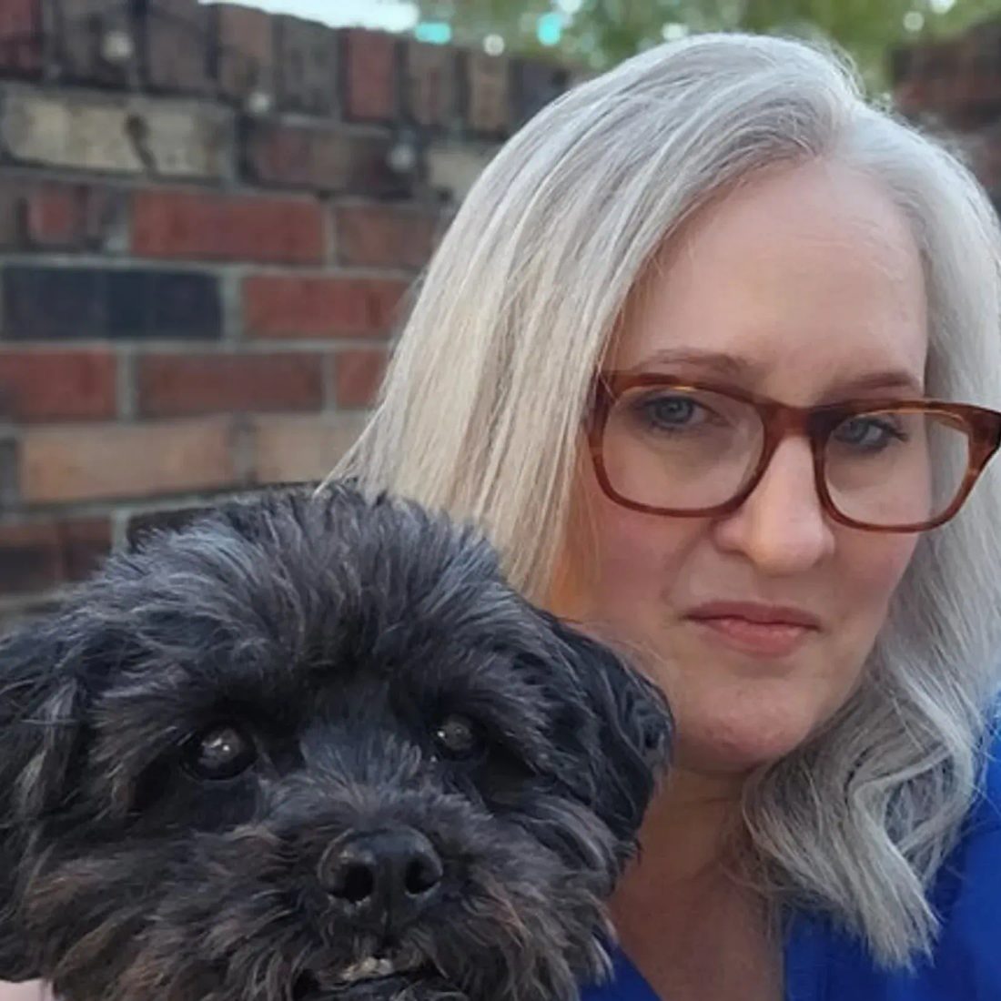Woman with glasses holding a small black dog in front of a brick wall